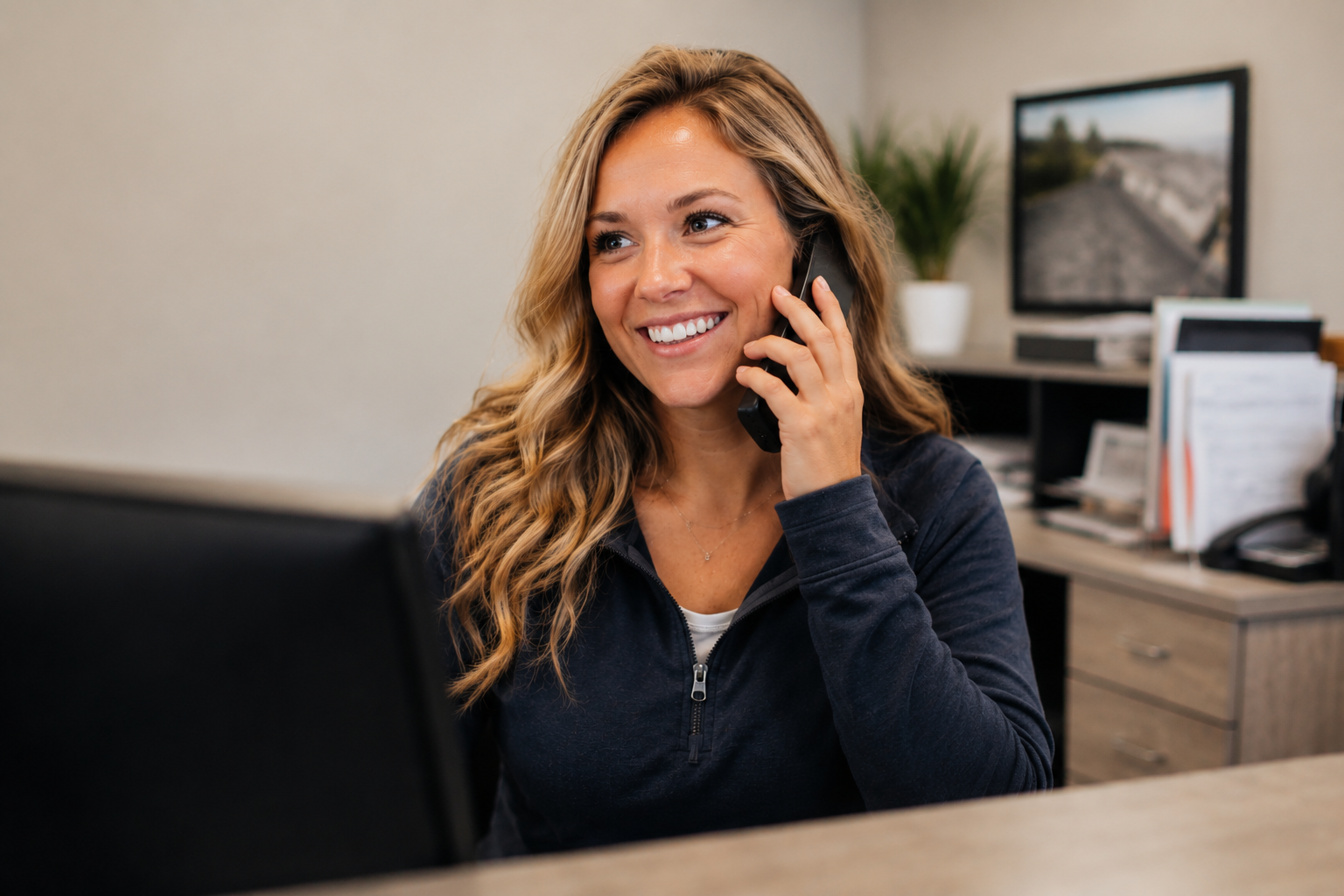 Smiling woman talking on a smartphone at a desk with a laptop and office background